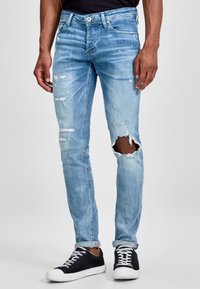 Light blue skinny jeans featuring distressed details and a large rip on the left knee, made from denim with a faded wash. Worn with black sneakers.
