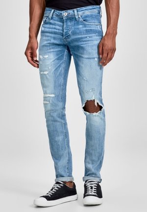 Jeans ajustados de color azul claro con detalles desgastados y un gran desgarro en la rodilla izquierda, fabricados en denim con un lavado descolorido. Llevados con zapatillas negras.