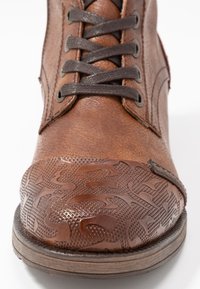 Brązowe skórzane buty z ciemnobrązowymi sznurówkami, z wzorzystym noskiem i teksturowanym wykończeniem górnej części, na białym tle.
