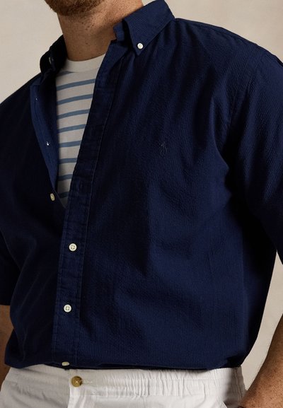 Un homme porte une chemise à boutons bleu marine sur un tee-shirt rayé gris, assortie à un short blanc. Ce style décontracté met en valeur une tenue estivale relaxante.