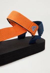 Narančasti platneni remeni s teksturiranom površinom, plavi detalji i oznaka s logotipom pričvršćeni na crnu pjenastu potplatu. Dizajn sandala, podesivog kroja.