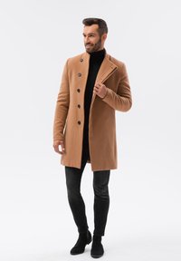 Cappotto in misto camel con una vestibilità su misura, colletto a bavero e cinque bottoni neri, abbinato a jeans slim neri e stivaletti.