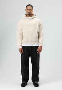 Beige hoodie met capuchon en een zak aan de voorkant, gecombineerd met zwarte broek met losse pasvorm. Gestructureerde stof, relaxte uitstraling en contrasterende witte lagen.