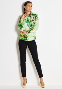 Vrouw in groene bloemetjes blouse met knopen, zwarte strakke broek en gele hakken, staand met hand nabij de kin tegen een witte achtergrond.