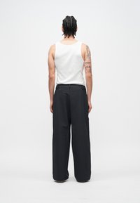 Balts bezroku topiņš, melnas platām kājām bikses ar gludu tekstūru un redzamām šuvēm. Tetovējums uz rokas ar tekstu, skatīts no aizmugures.