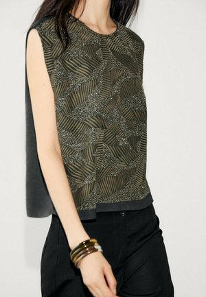 Sleeveless top with olive green and black geometric beadwork worn with black pants and stacked brown and gold bangles.
