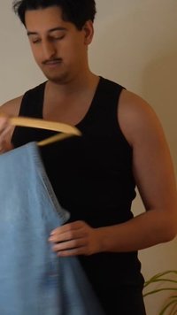 Mężczyzna w czarnej koszulce na ramiączkach trzyma jasnoniebieskie dżinsy na drewnianym wieszaku na tle gładkiej beżowej ściany.