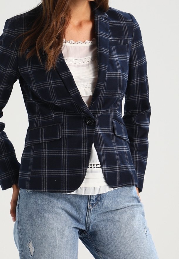 Femme portant un blazer à carreaux bleu marine sur un chemisier blanc orné de dentelle et un jean bleu clair déchiré, sur un fond uni.