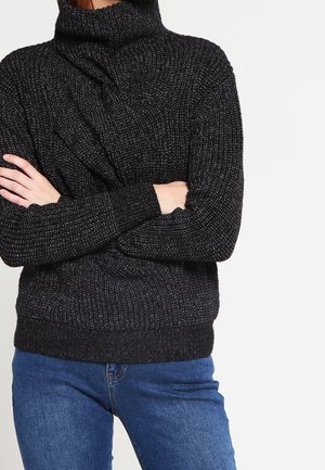 Personne portant un pull à col roulé noir en grosse maille avec les bras croisés et un jean bleu sur un fond blanc uni.