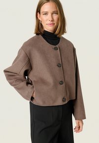 Brun cropped jakke lavet af blødt stof, med en rund halsudskæring, lange ærmer og fem sorte knapper foran.