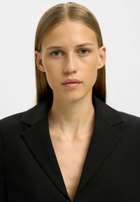 Černé sako s klopami a hlubokým výstřihem do V. Tkanina vypadá hladce s matným povrchem. Model má rovné, dlouhé vlasy.