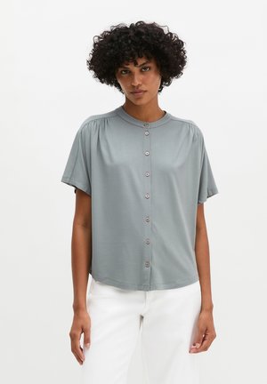 Femme aux cheveux bouclés portant un chemisier gris clair à manches courtes boutonné et un pantalon blanc, debout sur un fond blanc uni.