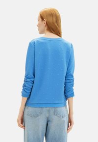Felpa blu con motivo a cuore testurizzato, colletto rotondo e maniche arricciate. Indossata con jeans azzurri, che mostrano una vestibilità comoda.