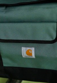 Sac en tissu vert et noir avec une petite étiquette blanche portant un logo Carhartt orange brodé sur le rabat de la poche avant.