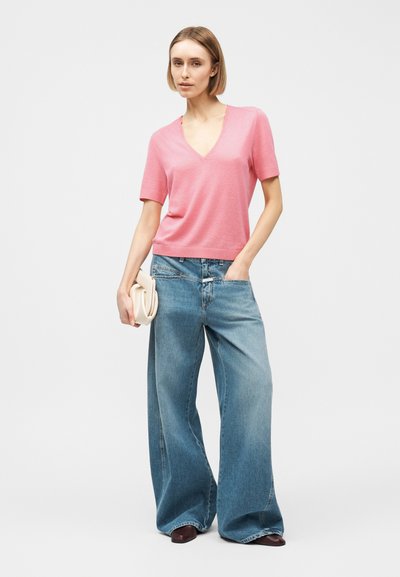 Femme portant un haut rose à manches courtes avec un col en V, un jean à jambes larges bleu, des chaussures foncées, tenant une petite pochette beige dans une main.