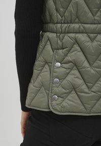 Quilted olivgrüne Weste mit Zickzackmuster, die eine taillierte Passform, silberne Druckknöpfe und einen schwarzen gerippten Ärmel aufweist.
