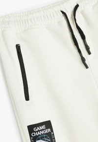 Pantalones de chándal blancos con una cintura elástica, cordones negros, bolsillos laterales con cremallera y una etiqueta negra con el texto "GAME CHANGER."