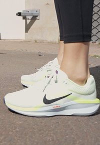 Bijeli Nike tenisice za trčanje s strukturiranim mrežastim gornjim dijelom, crnim swoosh logom i svijetlo žutom akcentom na potplatu. U kombinaciji s crnim tajicama.