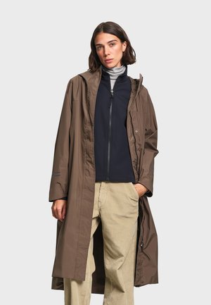 Femme portant un long manteau marron par-dessus une veste noire zippée, un col roulé gris et un pantalon beige en velours côtelé, debout avec une main dans la poche.