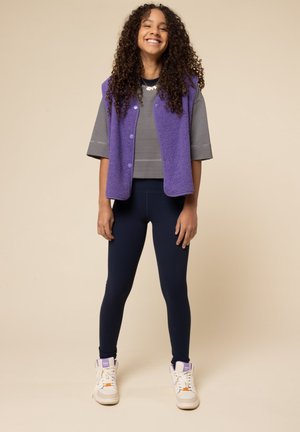 Paarse fleece vest over een grijs gestreept t-shirt met korte mouwen, gecombineerd met marineblauwe leggings en witte hoge sneakers met oranje en paarse accenten.
