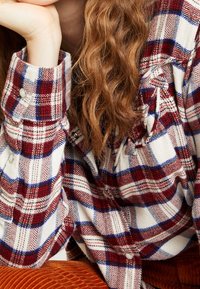 Rotes und blaues kariertes Flanellhemd mit weißem Grund, zeigt eine strukturierte Webart und Knopfdetails an Kragen und Manschetten.