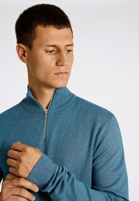 Blå stickad tröja med hög krage och kvartslång dragkedja. Ribbad mudd och fåll, slät textur med en subtil blandning av färger.