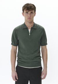 Polo de manga corta de color verde oscuro con cuello con cremallera, hecho de una tela de punto suave. Presenta mangas ajustadas y un corte relajado.