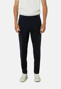 Marineblaue Hose im Slim-Fit, aus glattem Stoff gefertigt. Mit Reißverschluss, Gürtelschlaufen und Seitentaschen. Kombiniert mit weißen Sneakers.