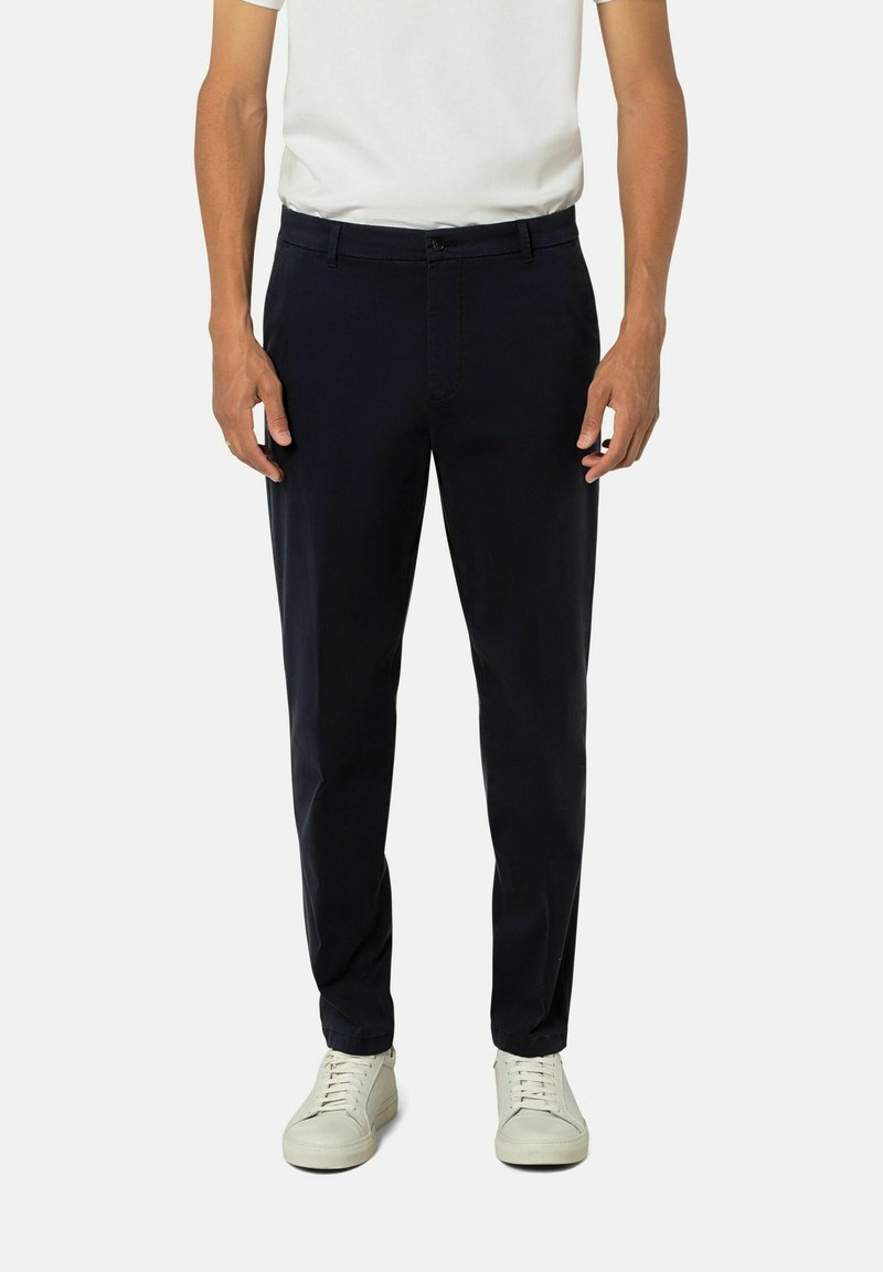 Marineblaue Hose im Slim-Fit, aus glattem Stoff gefertigt. Mit Reißverschluss, Gürtelschlaufen und Seitentaschen. Kombiniert mit weißen Sneakers.