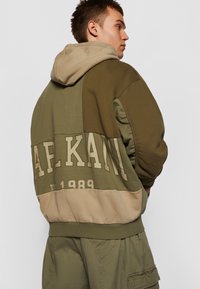 Kapucnis pulóver színblokk mintával olíva zöld és bézs színekben, hátul nagy, dombornyomott "A.P.KANI EST. 1989" felirattal.
