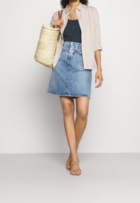 Gonna a trapezio in denim azzurro chiaro, con vita alta e chiusura a bottone, abbinata a una camicia beige con bottoni e una borsa in rafia.