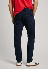 Dark blue denim jeans with a slim fit, featuring two back pockets and a black label at the waist, paired with white sneakers.