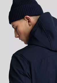 Felpa blu navy con colletto alto, caratterizzata da una texture liscia e dettagli di cucitura. Un berretto a coste scuro completa il look.