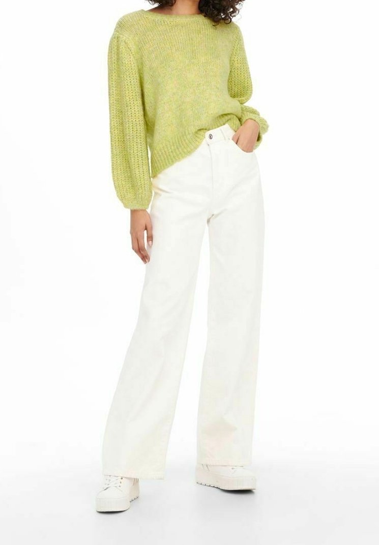 Personne portant un pull en maille vert clair à manches bouffantes, un pantalon blanc large et des chaussures blanches à plateforme, debout avec une main dans la poche.