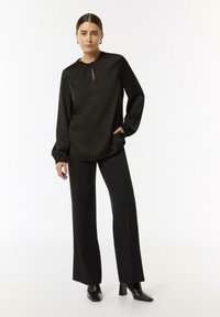 Schwarze Bluse mit langen Ärmeln und gerafftem Ausschnitt sowie einem Schlüsselaugen-Detail vorne, kombiniert mit schwarzen weiten Hosen und hohen Stiefeletten.