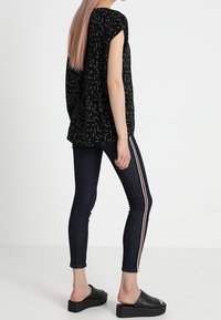 Person wearing black patterned short-sleeve top, dark skinny jeans with red and white side stripes, and black platform slip-on sandals, standing sideways.