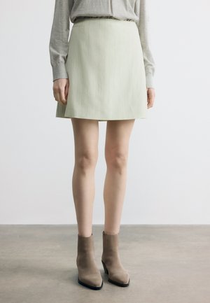 Vrouw die een beige mini-rok met krijtstreep draagt, gecombineerd met taupe enkellaarsjes, staand op een grijze vloer tegen een witte achtergrond.