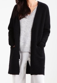 Dlouhý černý kardigan s otevřeným předním dílem, pružnými manžetami a kapsami. Vyroben z měkkého, strukturovaného materiálu, přeložený přes světle šedý svetr.