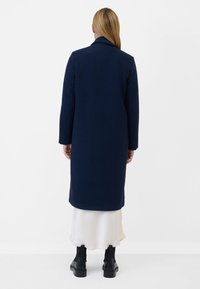 Mörkblå lång kappa med en strukturerad yta, med skarpt krage och långa ärmar, bärs över en krämfärgad kjol och svarta ankelstövlar.