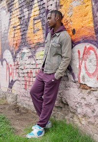 Giacca di jeans sopra un hoodie bordeaux e pantaloni jogger abbinati. Sneakers blu e bianche. Sullo sfondo, graffiti colorati su un muro di mattoni.