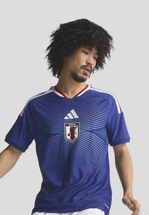 Blue soccer jersey with short sleeves, featuring white stripes, a V-neck, and a central crest with a bird design. Textured fabric with wavy lines.