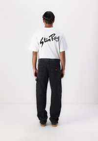 White short-sleeve t-shirt with black logo on back, paired with loose-fitting black cargo pants featuring multiple pockets and golden accents.