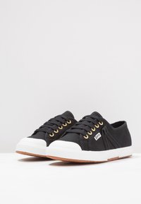 Schwarze Canvas-Sneaker mit weißen Gummisohlen, goldenen Ösen und strukturierten Oberflächenverzierungen. Mit runder Zehenform und niedrigem Schnitt.