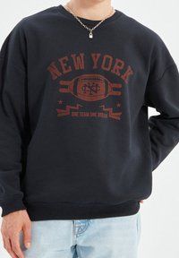Sudadera negra con impresión en rojo oscuro de "New York", que presenta un gráfico de fútbol y el eslogan "Un equipo, un sueño", con puños y dobladillo de canalé.
