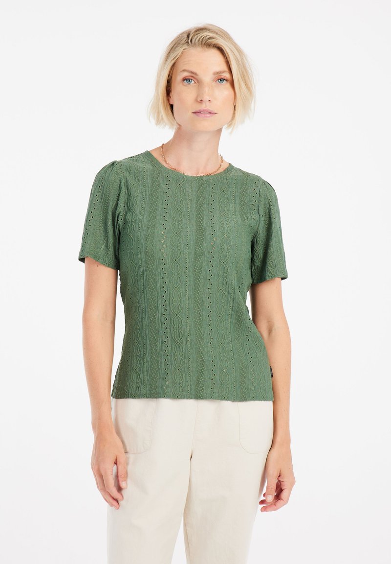 Top corto de manga corta verde con tejido texturizado y patrón de encaje. Corte recto y cuello redondo. Combinado con pantalones de color claro.
