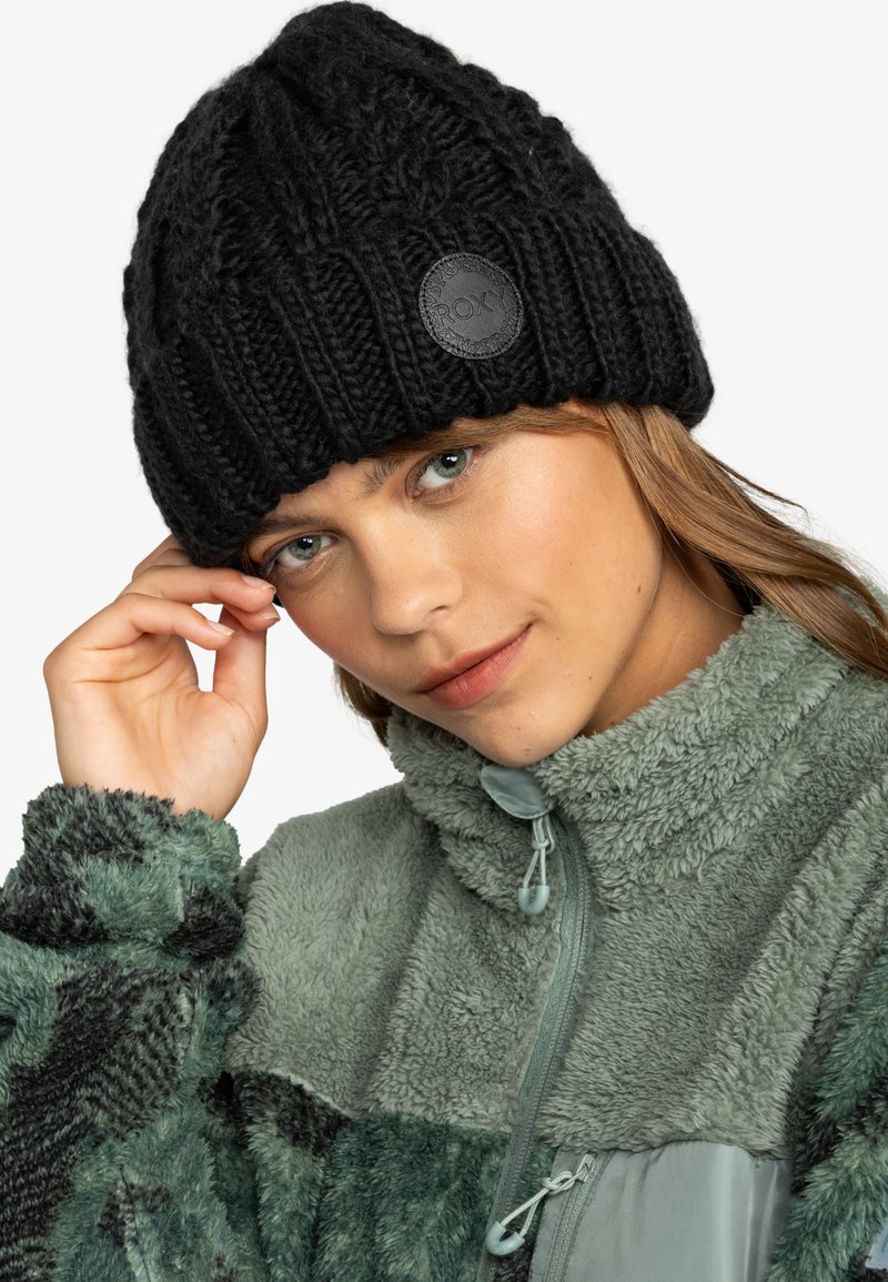 Black knitted beanie with a ribbed design and a circular, embossed Roxy logo. Model wears a green fleece jacket, showcasing textured fabrics.