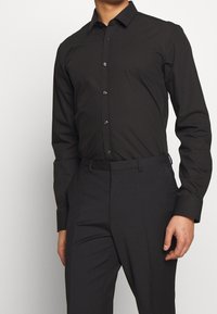 Czarna koszula z długim rękawem wykonana z gładkiej tkaniny, z klasycznym kołnierzykiem, plisą na guziki i dopasowanym krojem, zestawiona z ciemnymi spodniami.
