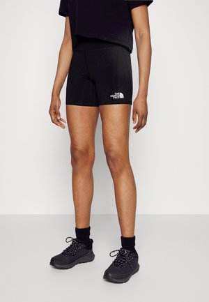 Osoba ubrana w czarne spodenki North Face, czarne skarpetki za kostkę oraz czarne buty sportowe stoi na jasnym tle.
