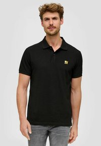 Schwarzes Poloshirt aus Baumwolle mit Kragen, kurzen Ärmeln und einem kleinen gelben Logo auf der linken Brust. Das Modell trägt graue Jeans.