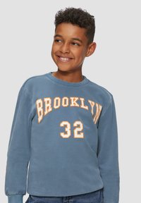 Glunderende jongen met kort, krullend haar in een blauwe sweatshirt met "BROOKLYN 32" in oranje en witte letters, staande tegen een effen achtergrond.